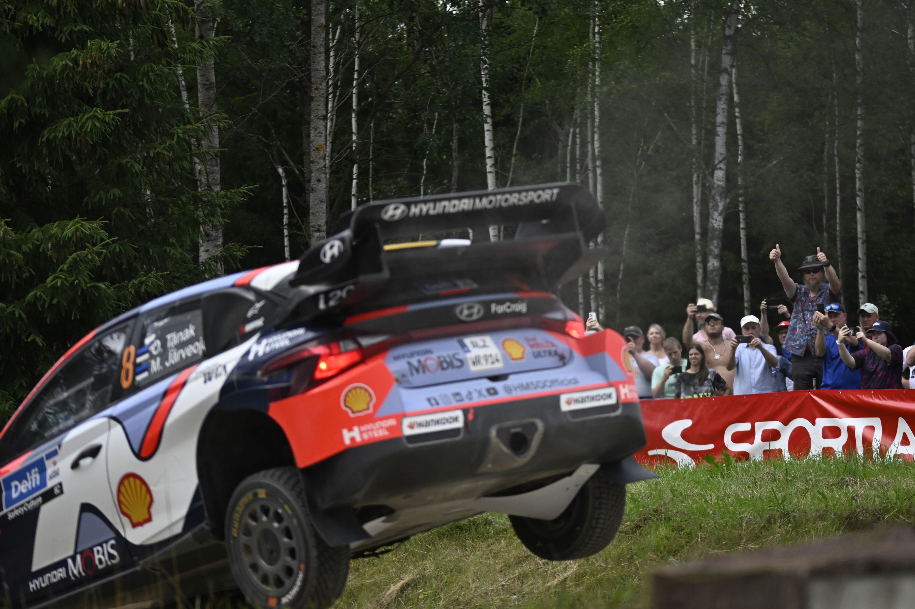 Ott Tänaku ja Martin Järveoja Hyundai i20 ralliauto õhus Truuta rallipargis hüppel. Taustal ergutavad pealtvaatajad, kes on kogunenud raja äärde Delfi Rally Estonia 2025 kiiruskatset jälgima.